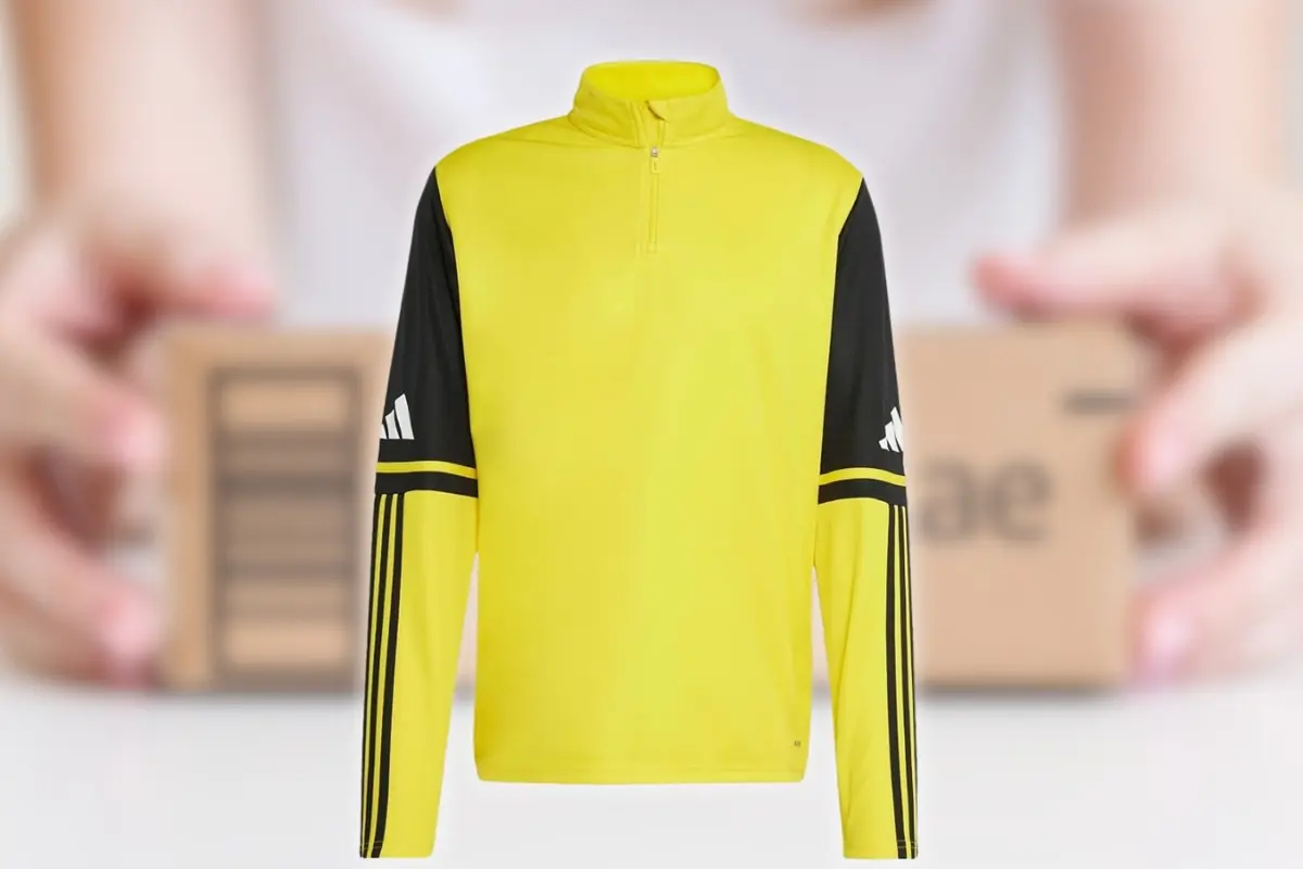 Sudadera deportiva amarilla de manga larga con detalles negros y cremallera corta al frente sobre fondo desenfocado de manos sosteniendo cajas de cartón