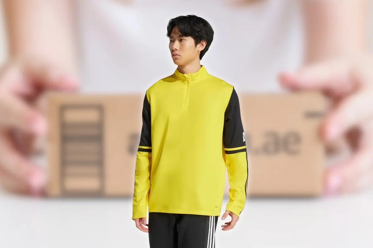 Hombre joven con sudadera deportiva amarilla y mangas negras de pie frente a un paquete de cartón sostenido por unas manos desenfocadas