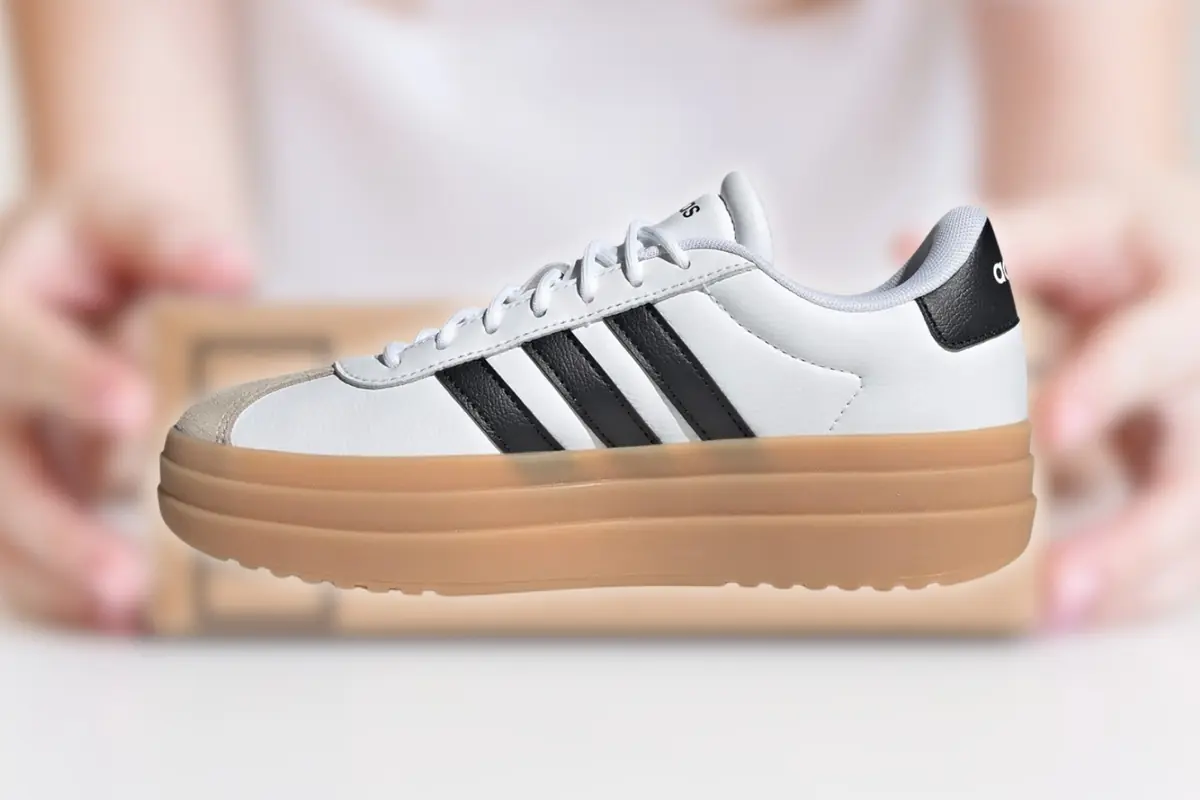 Zapatilla deportiva blanca con suela gruesa color goma y tres franjas negras sobre fondo desenfocado