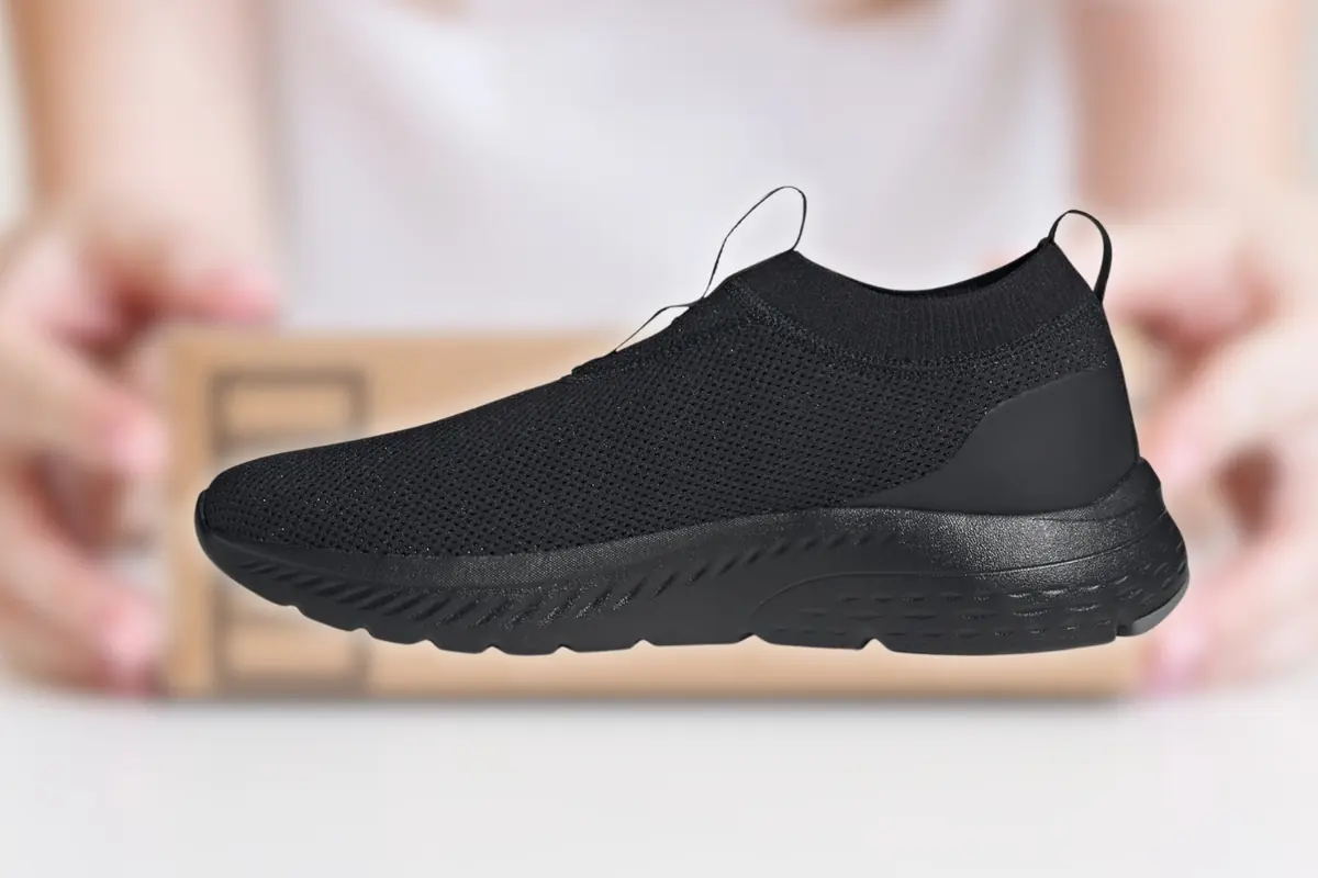 Zapatilla deportiva negra de malla ligera tipo slip on flotando frente a un fondo desenfocado con manos sosteniendo una caja de cartón