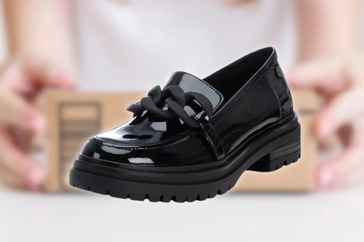 Mocasín negro de charol con suela gruesa y detalle de cadena sobre fondo desenfocado con caja de envío detrás