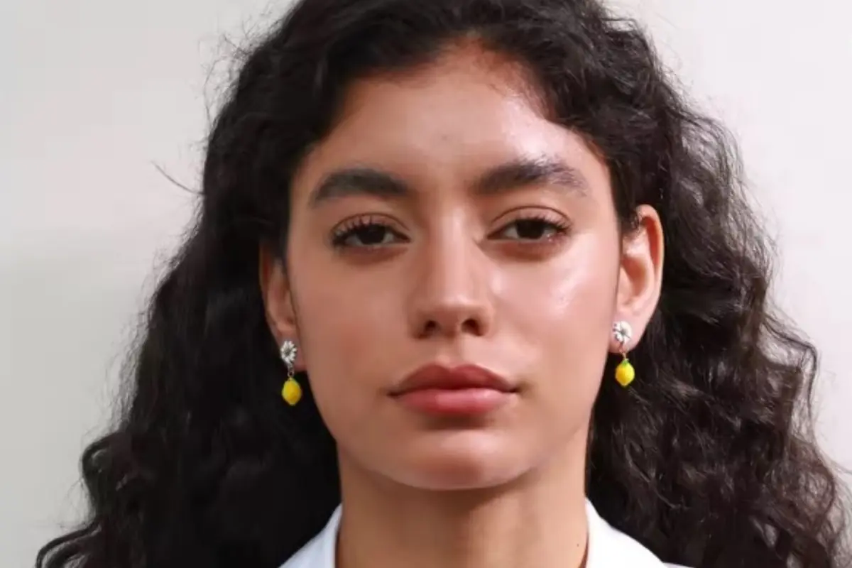 Mujer joven de cabello rizado oscuro mirando al frente con expresión seria y usando unos pendientes de limones