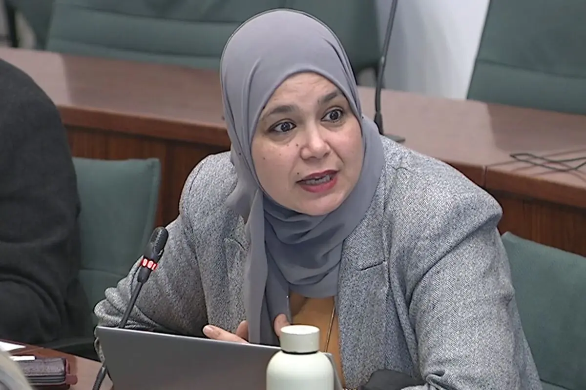 Mujer con hiyab gris hablando frente a un micrófono en una sala de reuniones oficiales mientras usa una computadora portátil