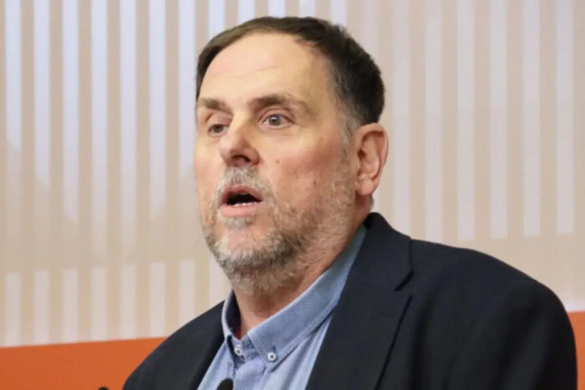 Home de mitjana edat amb barba encanosa parlant davant d’un micròfon i un fons amb franges verticals clares