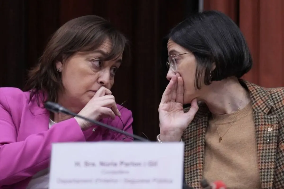 Dues dones assegudes en una taula de reunió s’inclinen i es tapen la boca mentre es xiuxiuegen a l’orella en un entorn institucional