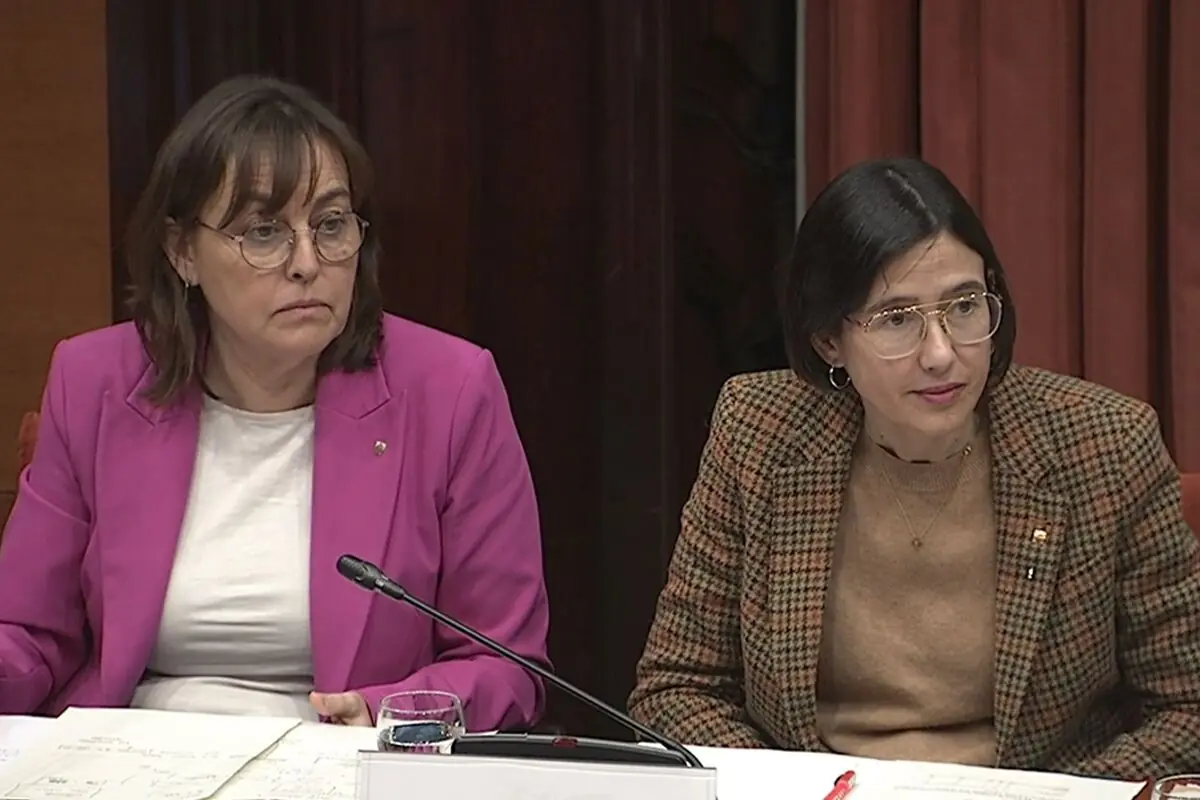 Dues dones assegudes en una taula durant una reunió formal amb un micròfon i documents al davant