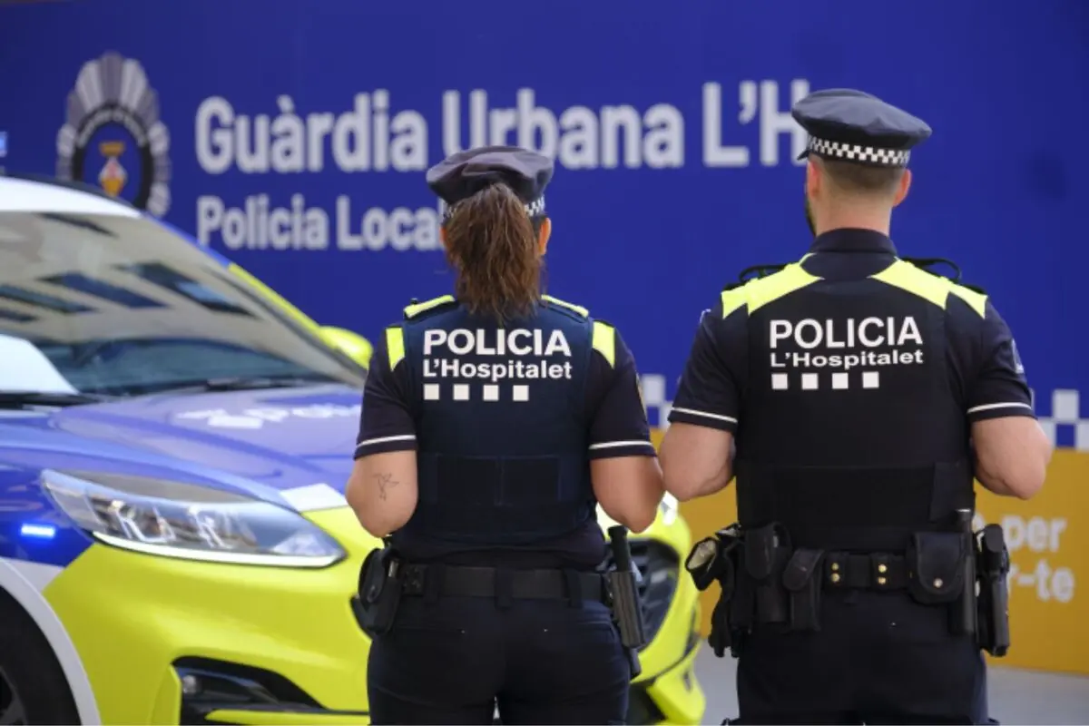 Dos agentes de la Policía Local de L’Hospitalet de espaldas frente a un coche patrulla y un cartel de la Guàrdia Urbana