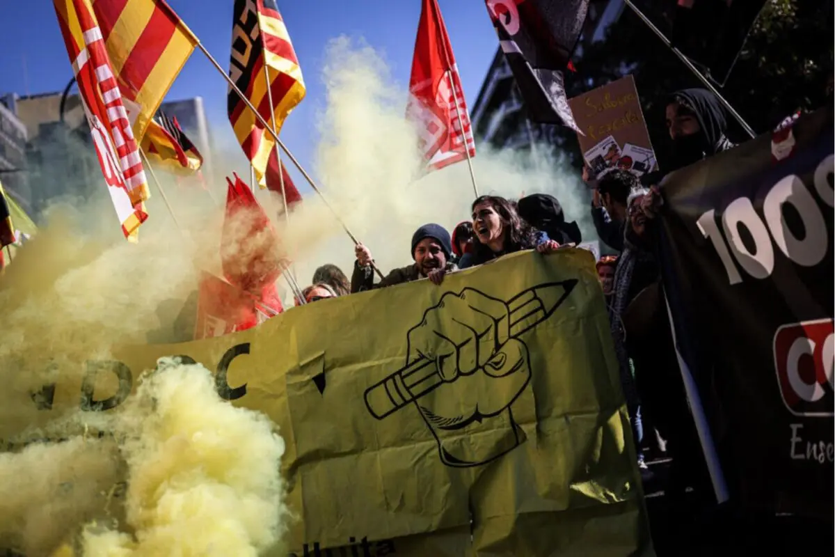 Manifestació amb persones sostenint pancartes i banderes de colors entre fum groc, destacant un gran cartell groc amb un puny alçat que agafa un llapis