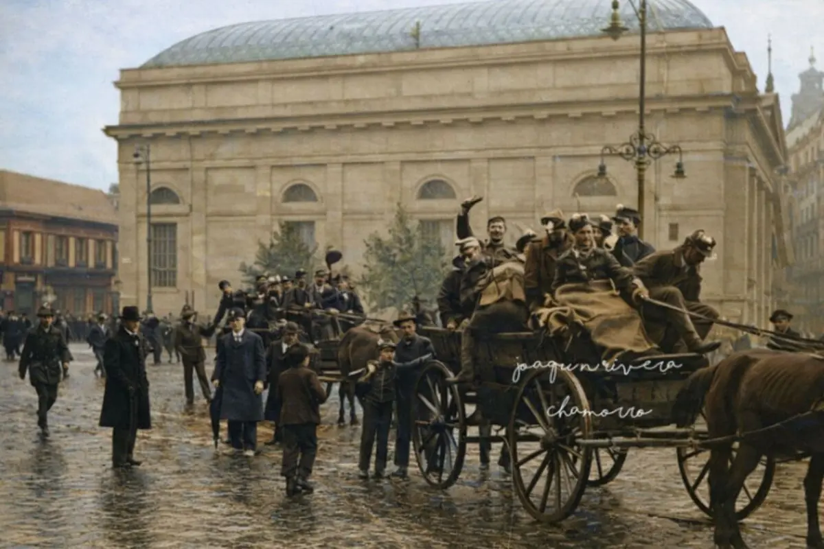 Carros tirats per cavalls plens de soldats i civils avancen per un carrer empedrat davant d’un gran edifici històric mentre la gent camina i observa l’escena; es tracta d’una imatge de la revolució dels Crisantems.