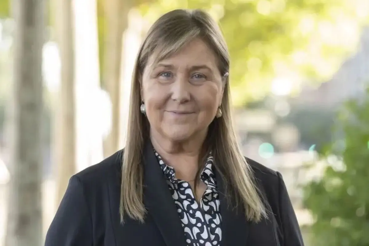Retrat d’una dona de mitjana edat amb jaqueta fosca i brusa estampada somrient a l’aire lliure amb un fons desenfocat d’arbres i llum natural