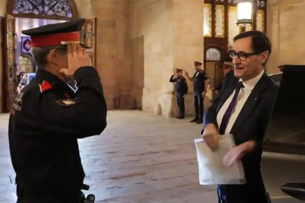 Un agent de policia uniformat saluda militarment un home amb vestit i ulleres que sosté uns documents a l’interior d’un edifici institucional