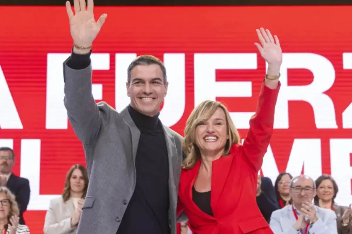Dos personas sonrientes levantan la mano saludando al público en un acto político con un gran cartel rojo de fondo y varias personas aplaudiendo detrás