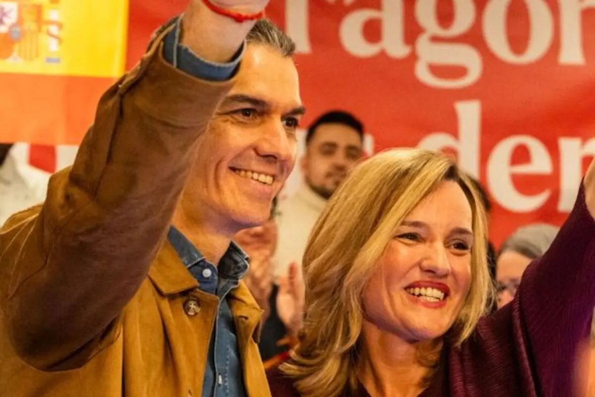 Dos personas sonrientes levantan el brazo en un acto público con una bandera de España y un cartel rojo de fondo Dos personas sonrientes levantan el brazo en un acto público con una bandera de España y un cartel rojo de fondo