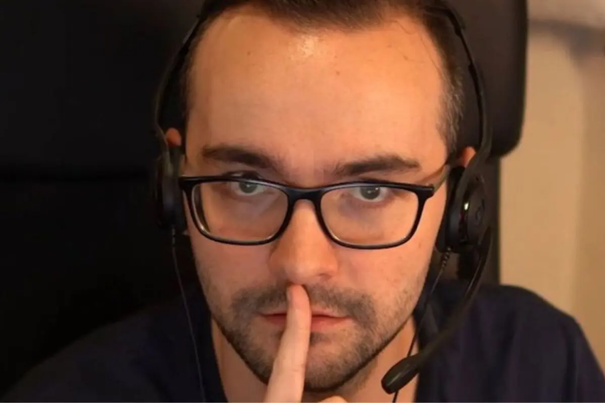 Home amb ulleres i auriculars mirant a la càmera mentre es porta un dit als llavis en gest de silenci