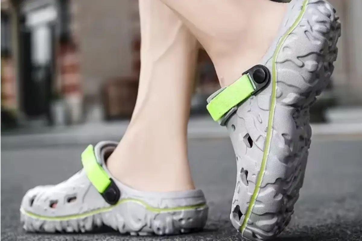 Zuecos deportivos grises con suela texturizada y correa verde fosforescente usados por una persona caminando sobre el asfalto