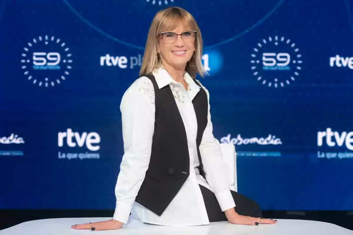 Gemma Nierga amb ulleres i cabell ros laci, vestida amb una camisa blanca i una armilla negra, està asseguda en una taula davant d'un fons blau amb logotips de RTVE i 