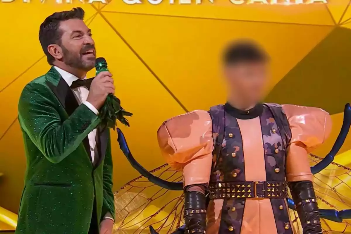 Arturo Valls amb un vestit verd brillant sosté un micròfon mentre és al costat d'una persona disfressada amb un vestit colorit i voluminós de Mosca en un escenari il·luminat a Mask Singer.