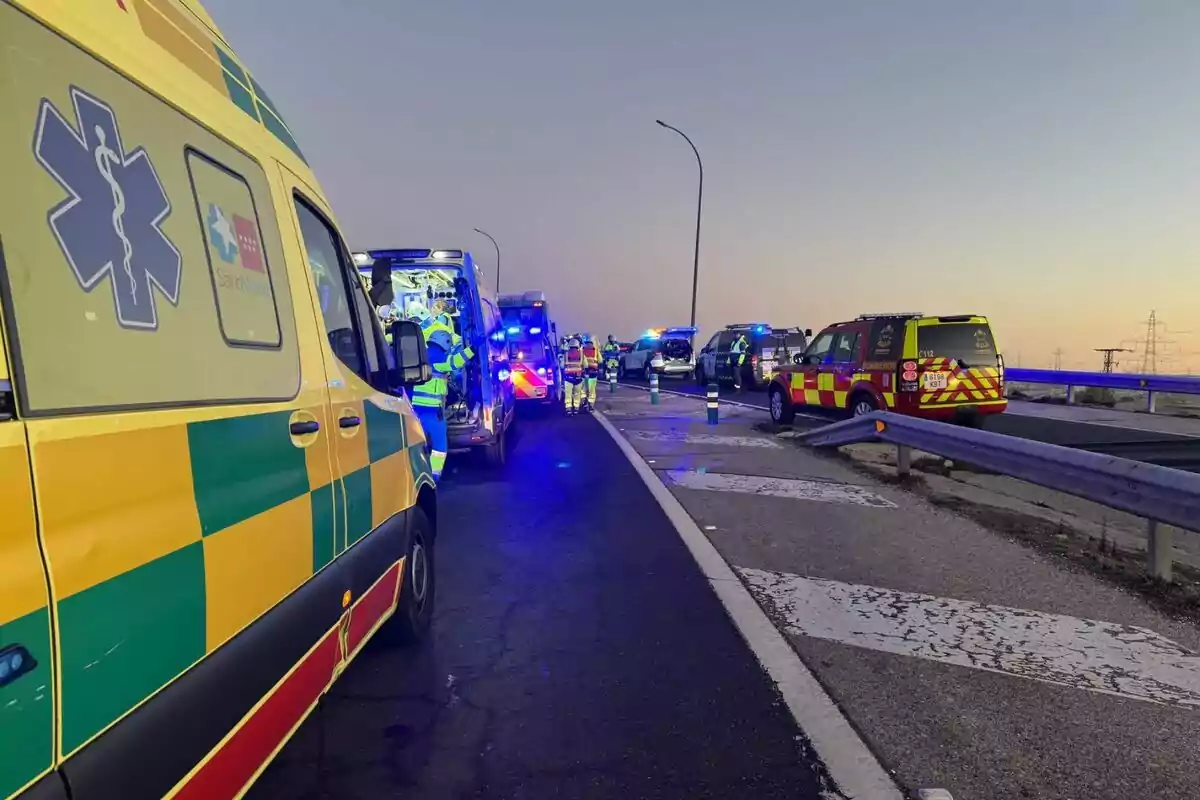 Varias ambulancias y vehículos de emergencia están estacionados en una carretera al atardecer con personal de emergencia trabajando en el lugar.