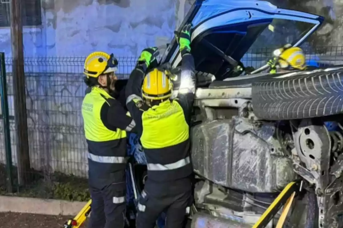 Dos bombers amb cascos grocs i armilles reflectores treballen en un vehicle bolcat.