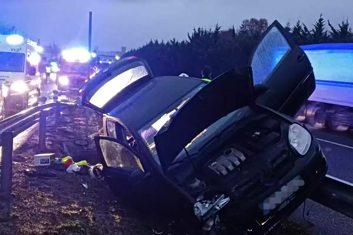 Un cotxe accidentat a la vora d?una carretera amb llums d?emergència de vehicles de rescat al fons.