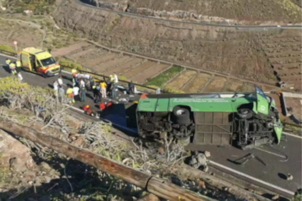 Un autobús verd bolcat en una carretera amb una ambulància i diverses persones al voltant en un entorn muntanyós.