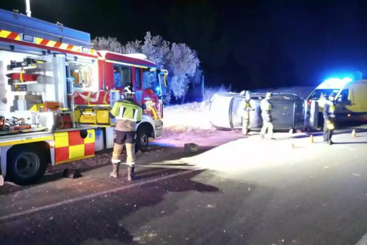 Un camió de bombers i una ambulància a l'escena d'un accident de trànsit amb un cotxe bolcat a la carretera mentre els bombers treballen al lloc.