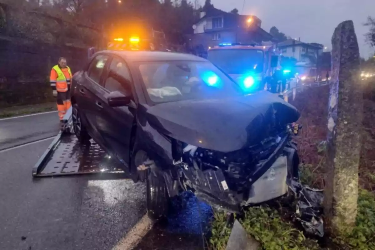 Un cotxe negre amb la part davantera danyada està sent remolcat per una grua en una carretera mullada mentre un treballador amb armilla reflectora observa l'escena i hi ha llums d'emergència encesos al fons.