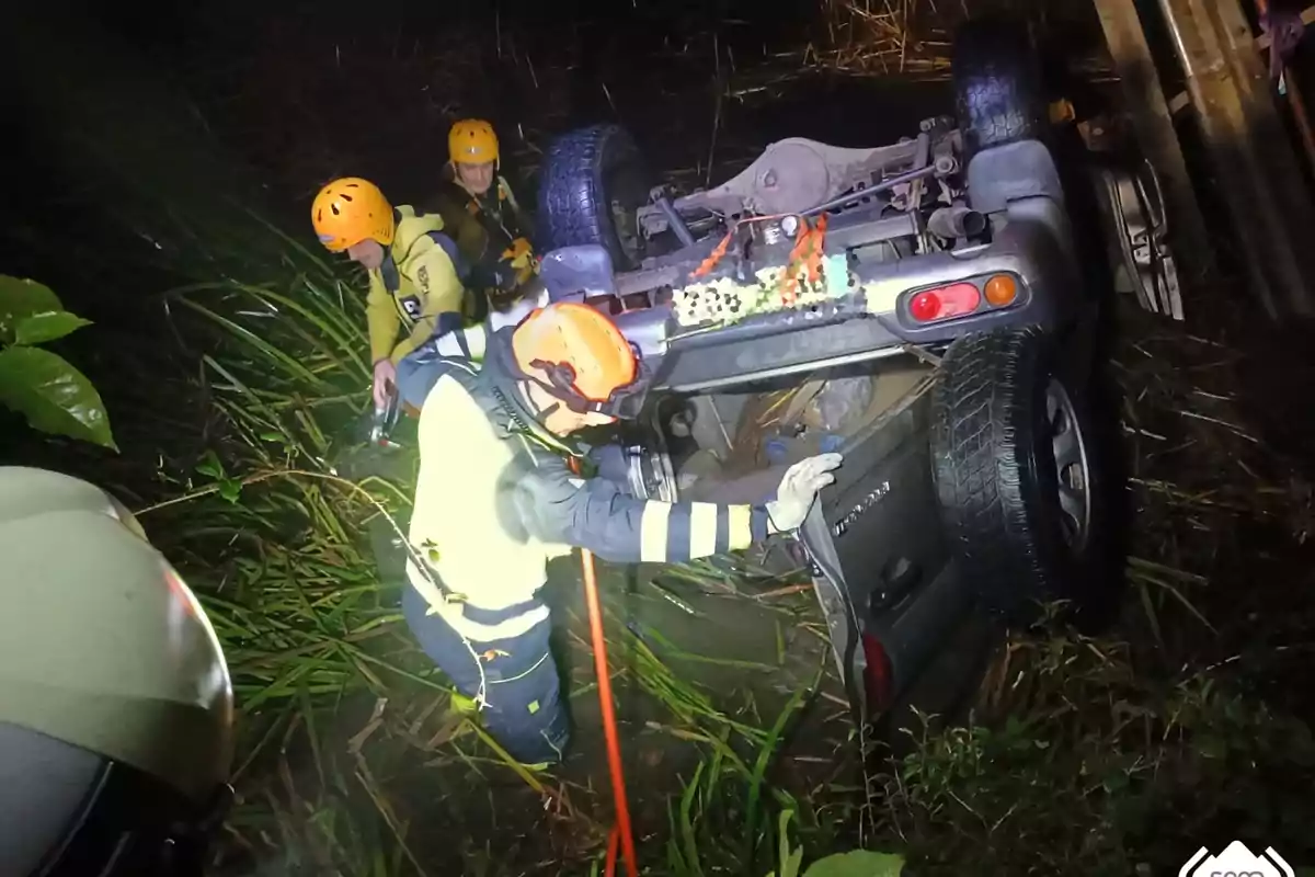 Rescatistes amb cascos i equip de seguretat treballen al lloc d'un accident on un vehicle està bolcat sobre el sostre en una àrea amb vegetació.