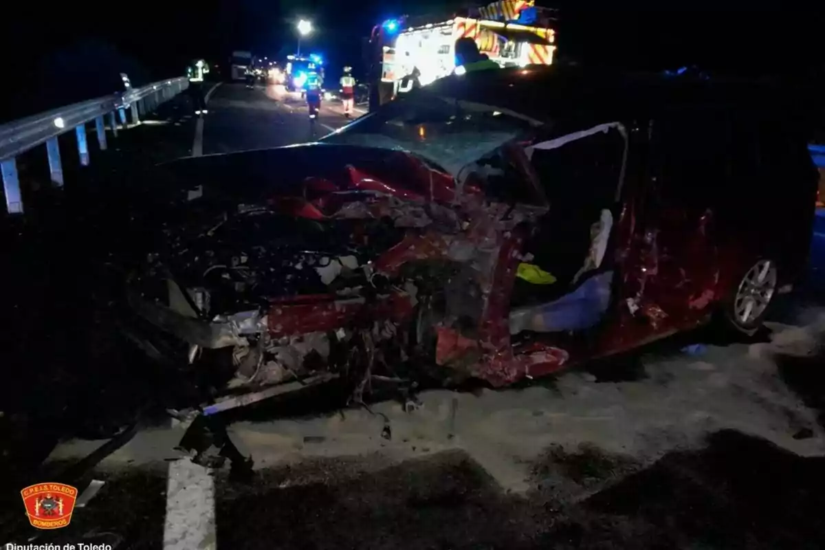 Un cotxe vermell greument malmès en un accident nocturn a una carretera amb presència de bombers i vehicles d'emergència il·luminats al fons.