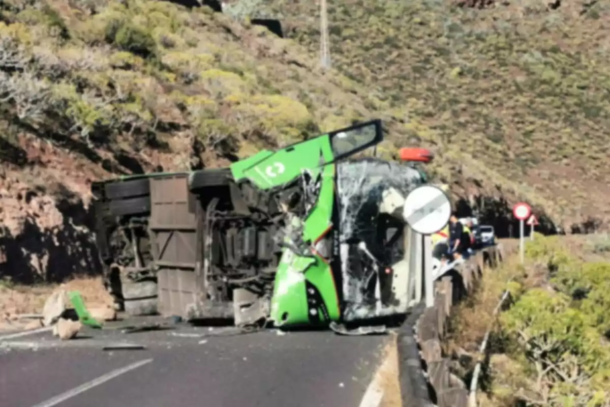Un autobús verd bolcat en una carretera de muntanya amb danys visibles al vehicle i alguns cotxes i persones al fons.
