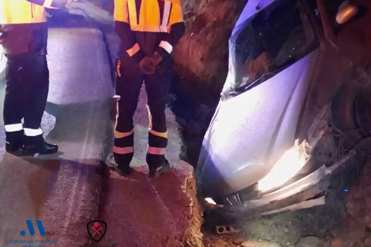 Un cotxe es troba enfonsat parcialment en una rasa al costat d'una carretera mentre dues persones amb uniformes de seguretat observen l'escena.