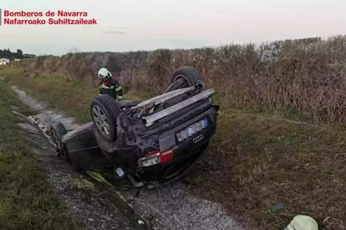 Un cotxe bolcat en una rasa al costat d´un camí rural amb un bomber de peu a prop del vehicle.