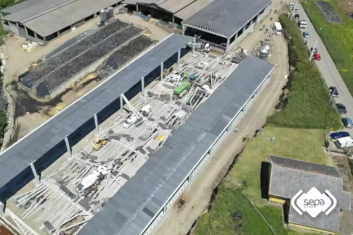 Vista aèria d'una nau industrial en construcció amb maquinària i materials a l'interior, envoltada d'altres edificis i una carretera amb diversos vehicles estacionats. Vista aèria d'una nau industrial en construcció amb maquinària i materials a l'interior, envoltada d'altres edificis i una carretera amb diversos vehicles estacionats.