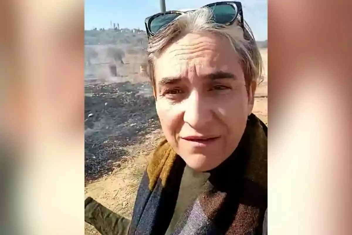 Una persona amb ulleres de sol al cap és a l'aire lliure en una àrea amb vegetació cremada i fum al fons.