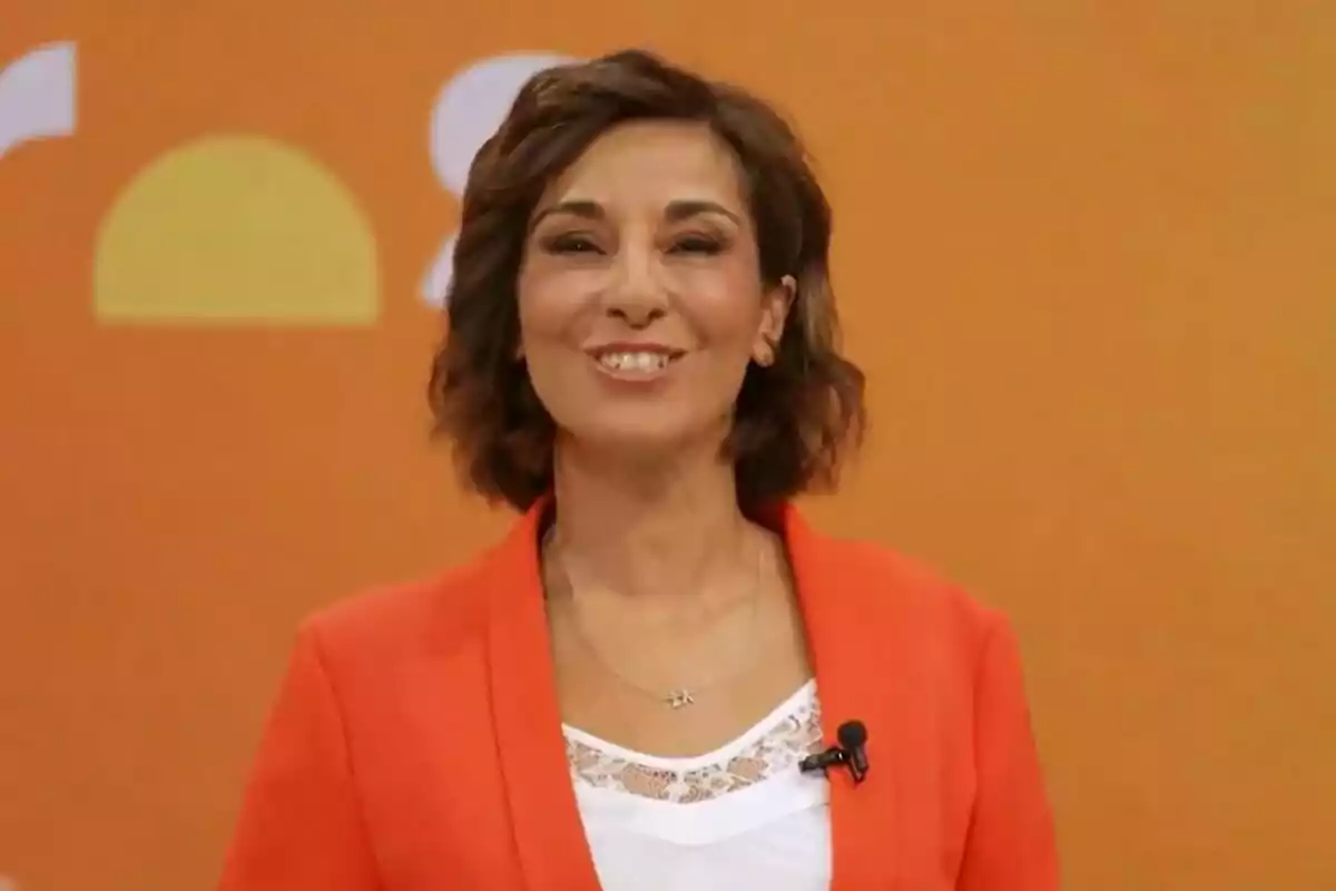 Captura de Adela González con cabello corto y ondulado, vestida con un blazer naranja y una blusa blanca, sonríe frente a un fondo naranja con un micrófono de solapa en la promo de Mañaneros