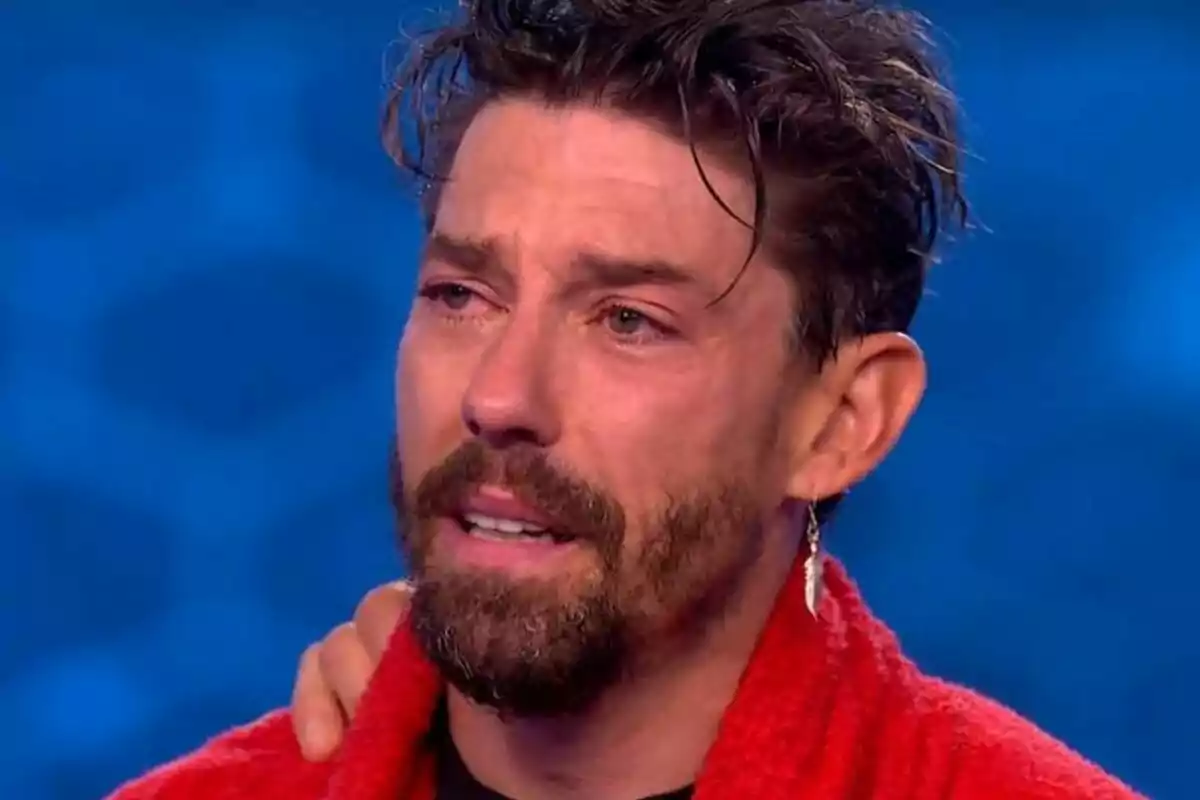 Adrián Lastra con barba y cabello oscuro parece emocionado mientras lleva una bata roja y un pendiente en la oreja en El Desafío.