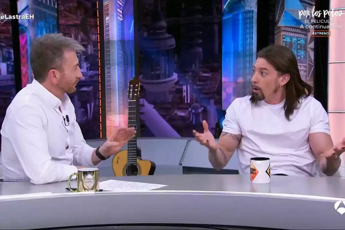 Pablo Motos i Adrián Lastra conversen a El Hormiguero, un d'ells gesticula amb les mans mentre l'altre escolta atentament; hi ha una guitarra al fons i tasses sobre la taula. Pablo Motos i Adrián Lastra conversen a El Hormiguero, un d'ells gesticula amb les mans mentre l'altre escolta atentament; hi ha una guitarra al fons i tasses sobre la taula.