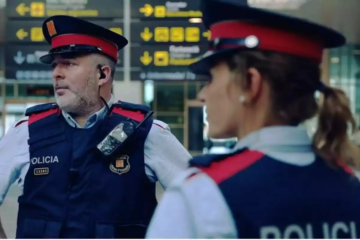 Dos agents de policia amb uniforme i gorra estan drets en un aeroport, un porta un dispositiu de comunicació a l'armilla.