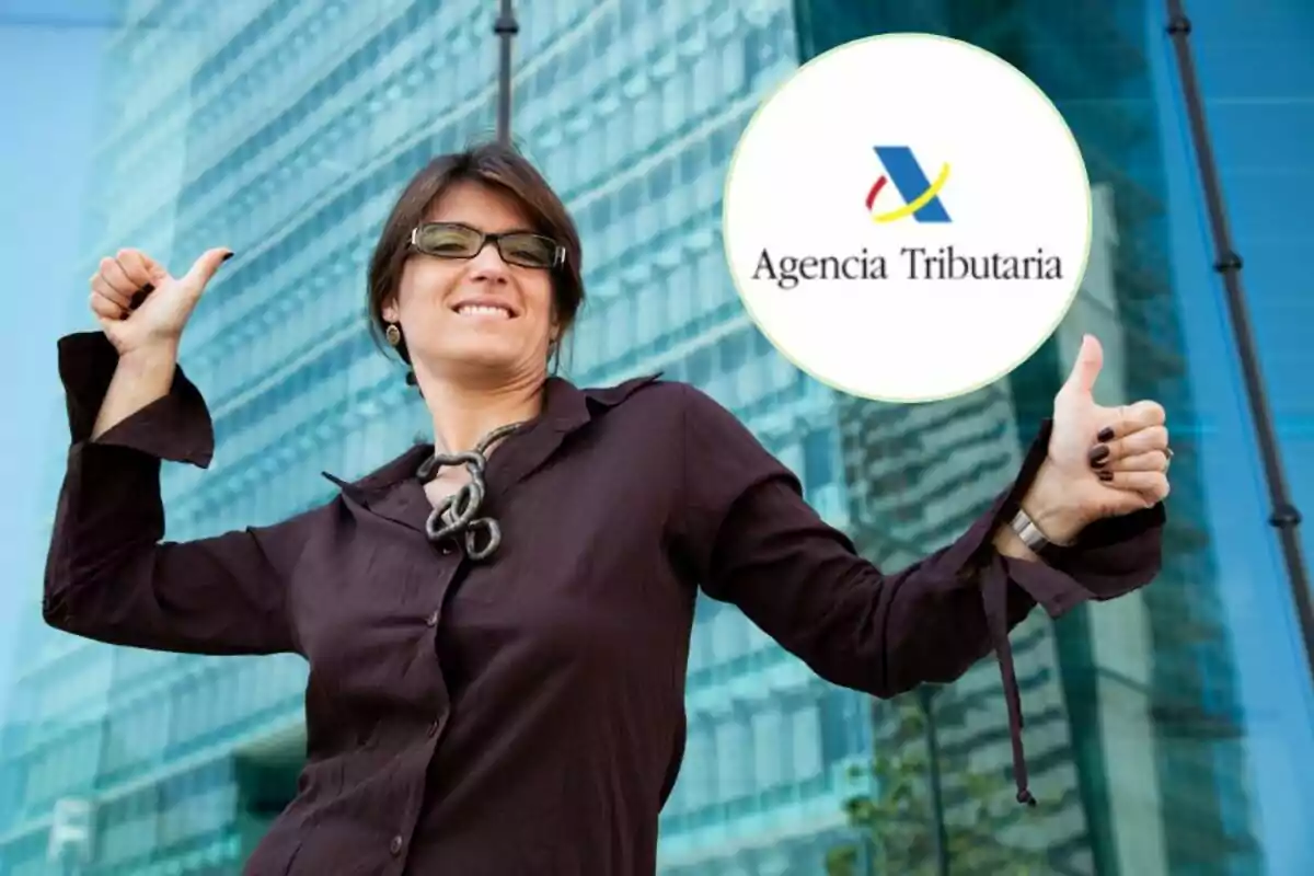 Mujer sonriendo con pulgares hacia arriba frente a un edificio moderno con el logo de la Agencia Tributaria.