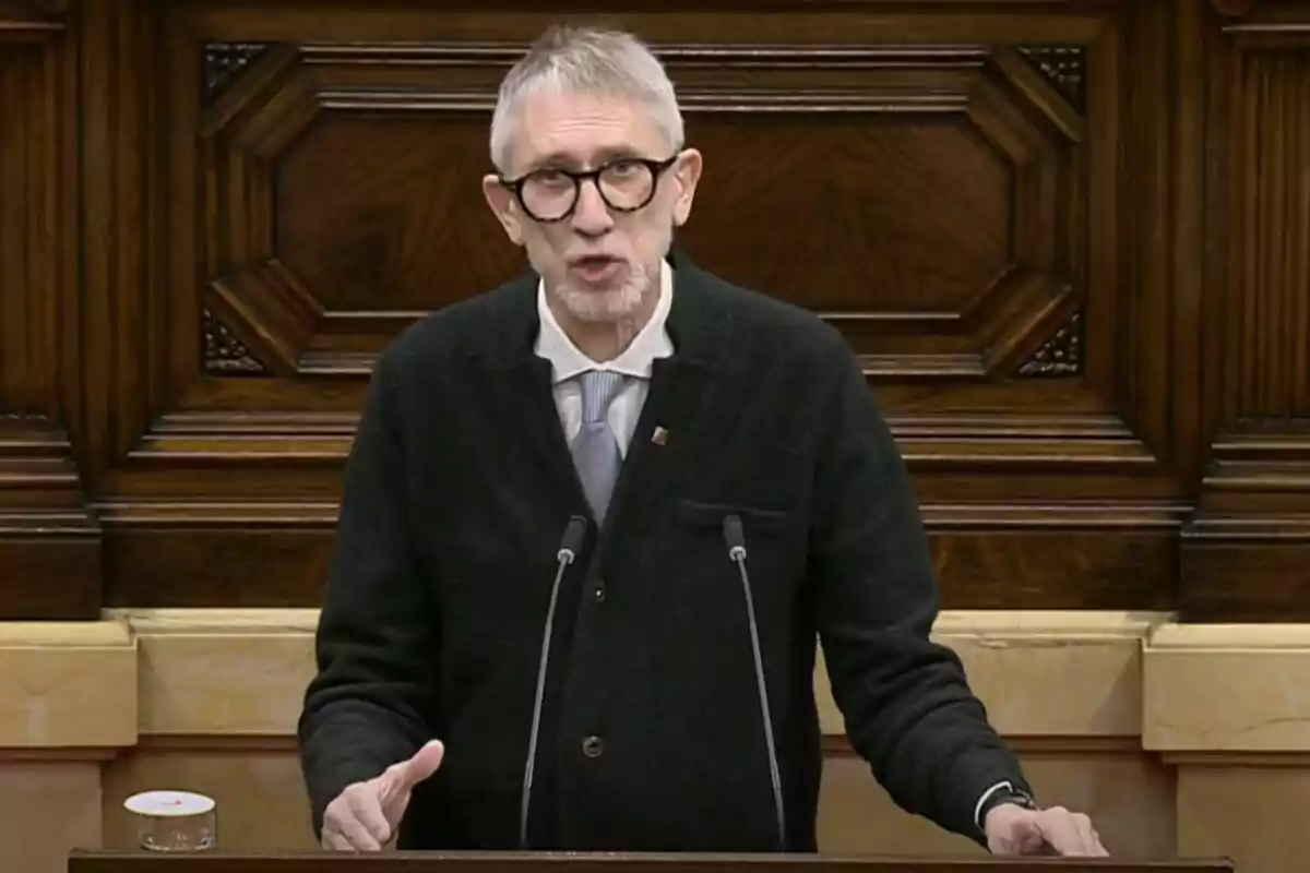 A man with glasses and a beard speaking at a wooden podium.