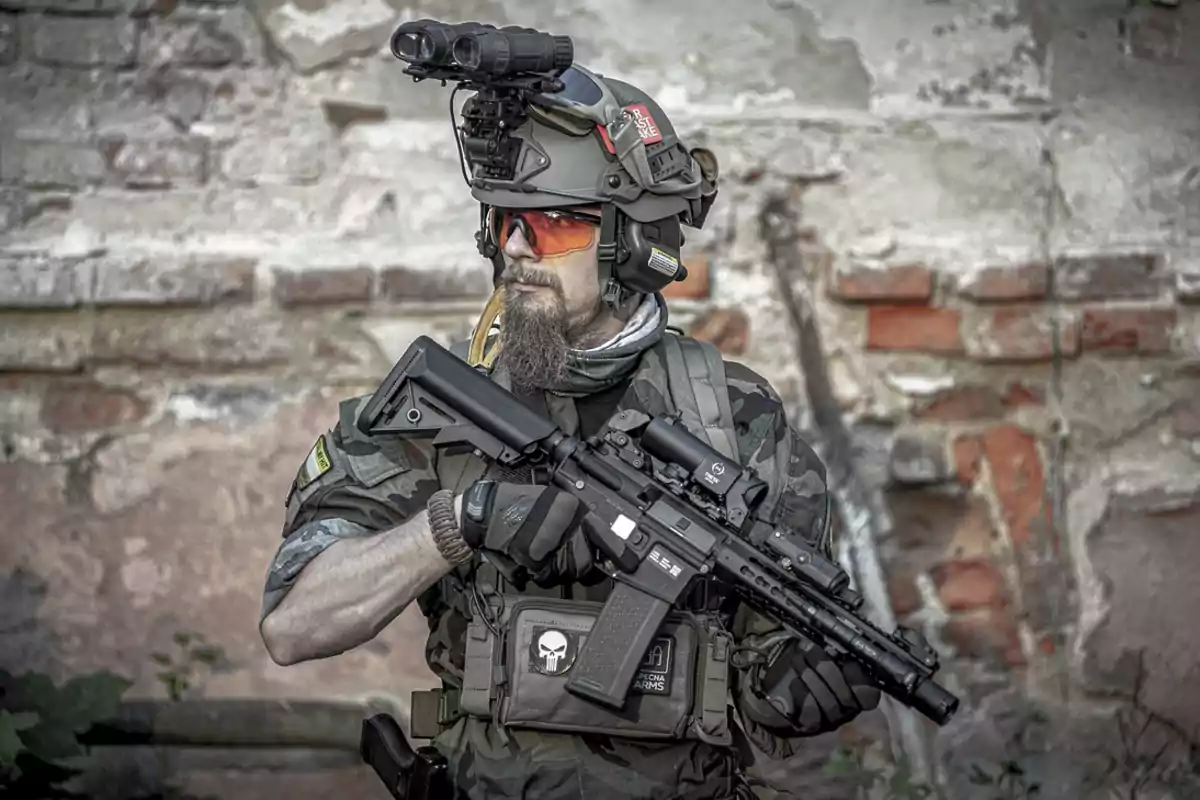 Man with a beard and orange glasses wearing a military uniform, tactical helmet, and holding a rifle in front of a weathered brick wall