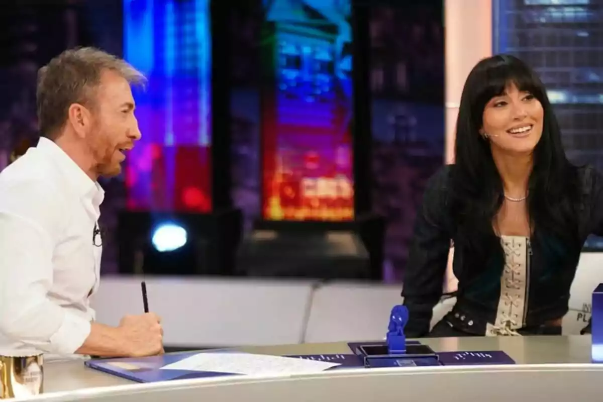 Aitana i Pablo Motos conversen i somriuen a 'La Revuelta' amb llums de colors al fons i papers sobre la taula. Aitana i Pablo Motos conversen i somriuen a 'La Revuelta' amb llums de colors al fons i papers sobre la taula.