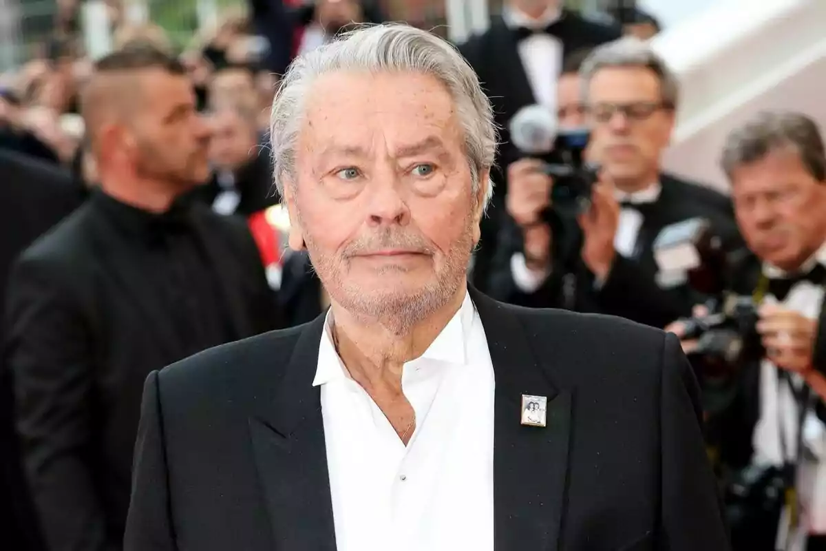 Alain Delon amb cabell canós i barba curta, vestit amb un vestit negre i camisa blanca, en un esdeveniment formal amb diverses persones i fotògrafs al fons. Alain Delon amb cabell canós i barba curta, vestit amb un vestit negre i camisa blanca, en un esdeveniment formal amb diverses persones i fotògrafs al fons.