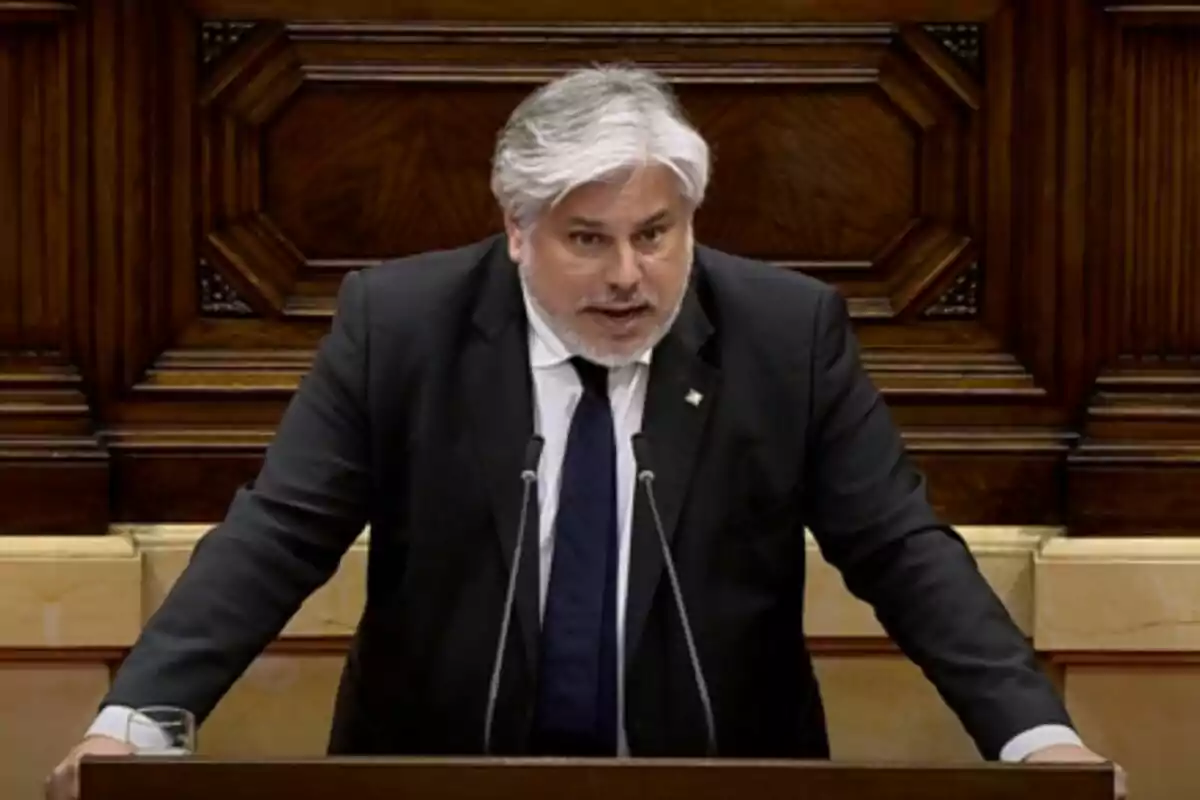 Un home de cabells canosos i barba està parlant en un faristol de fusta en un entorn formal.