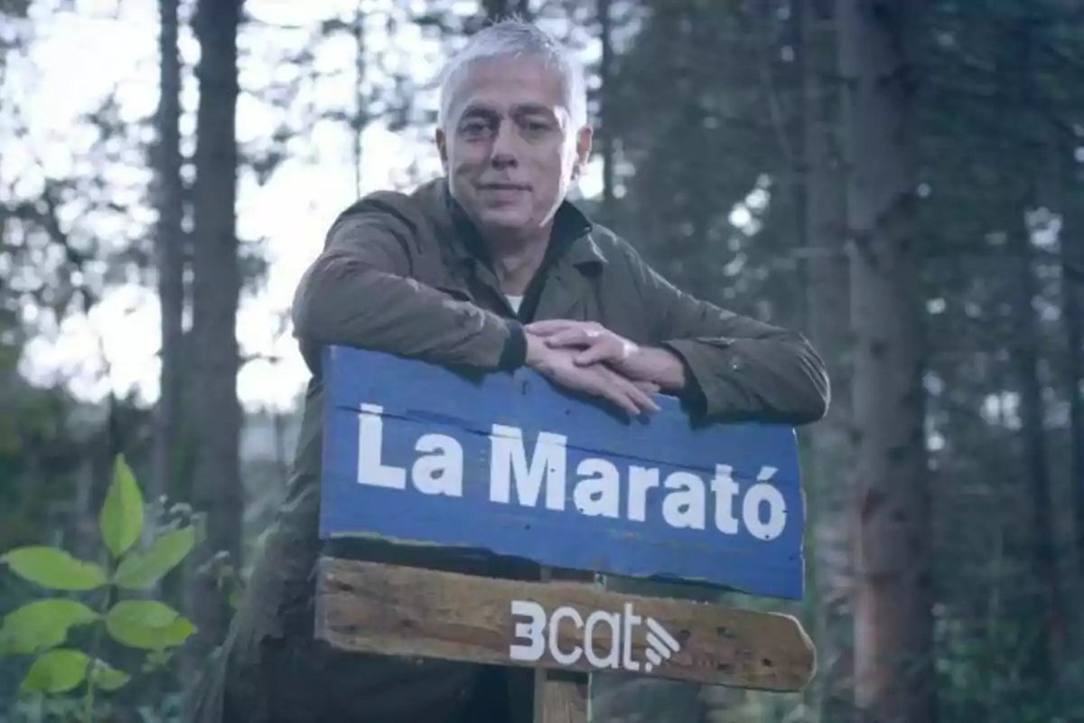 Albert Om de cabell canós recolza els seus braços sobre un cartell blau que diu “La Marató” en un entorn boscós.