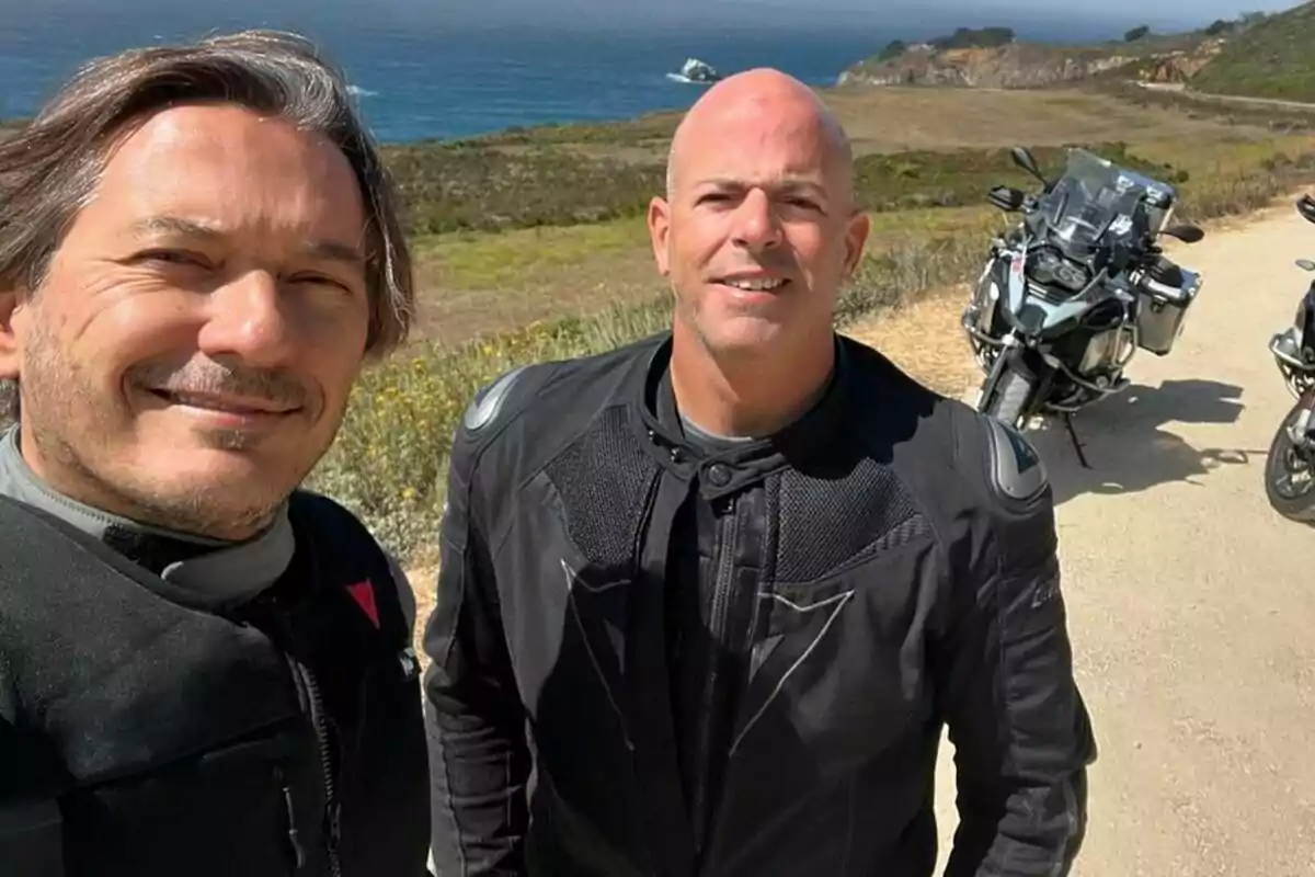 Dos homes amb jaquetes de motociclista posen per a una selfie exterior amb motocicletes i un paisatge costaner de fons.