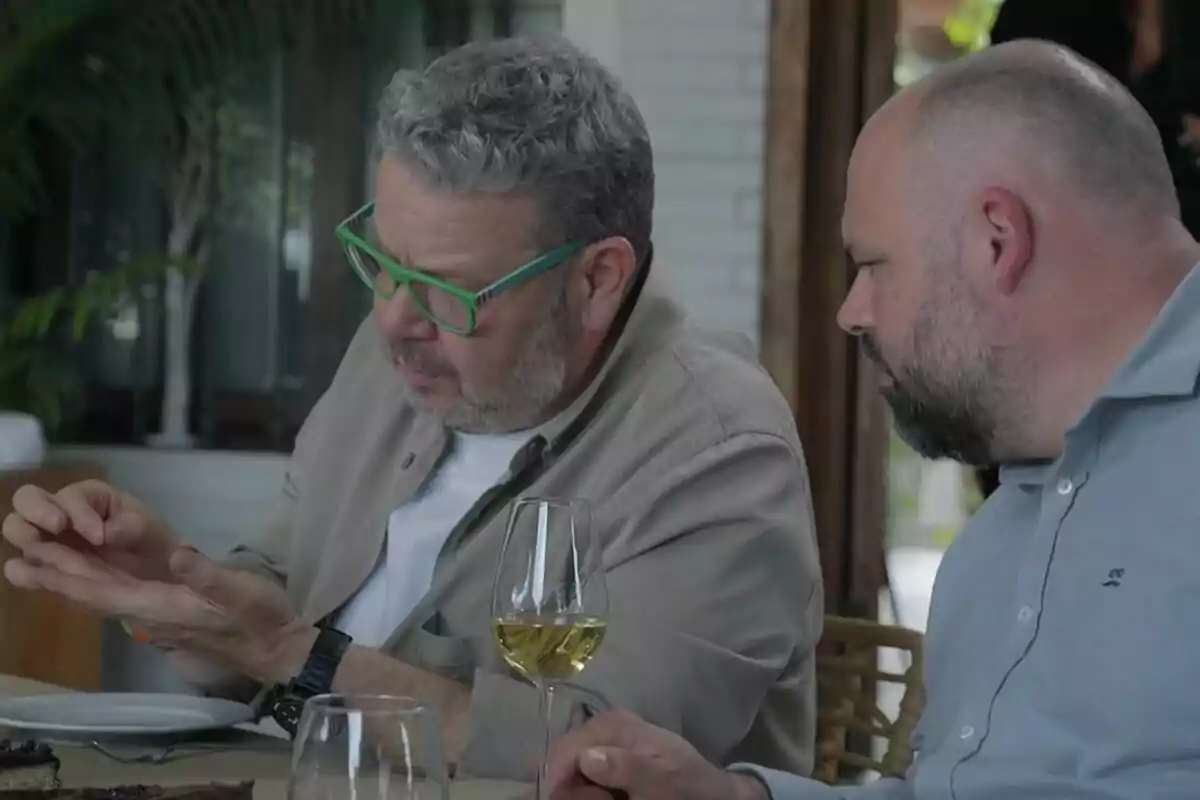 Alberto Chicote mirando un pelo en un plato junto a otro hombre mientras conversan en una mesa con copas de vino y comida en Batalla de Restaurantes.