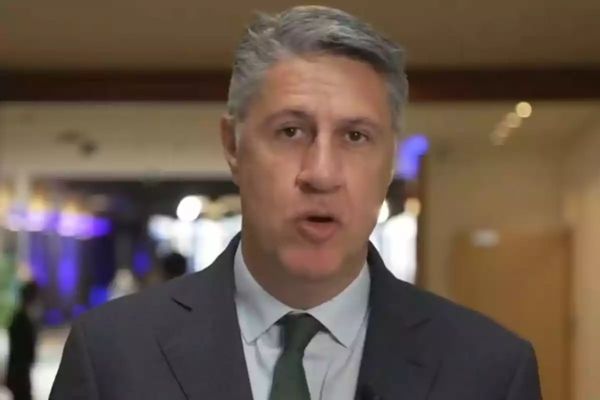 Home de cabell canós amb vestit i corbata verda parlant davant de la càmera en un entorn interior.