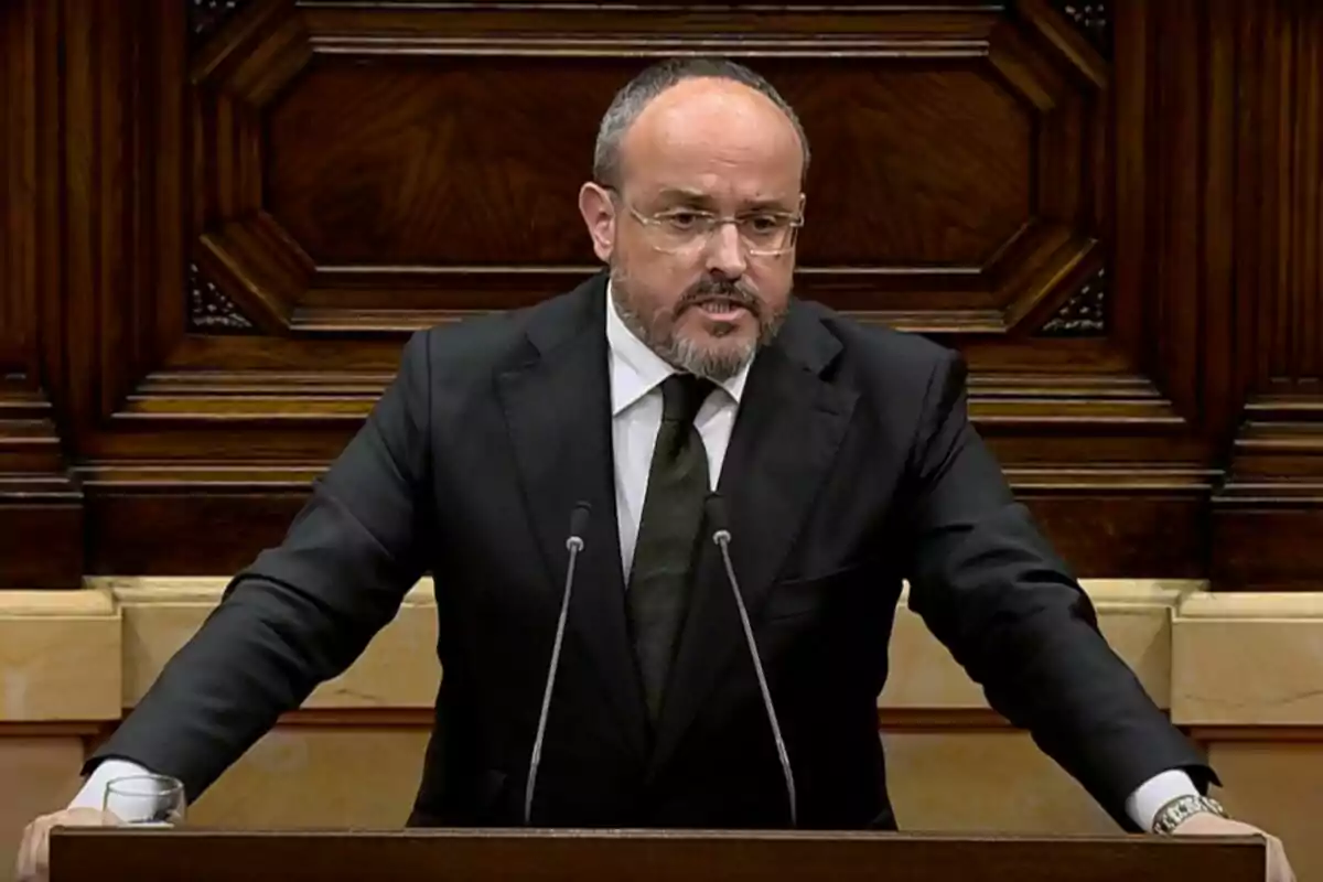 Un home amb barba i ulleres està parlant en un faristol de fusta amb un fons decoratiu. Un home amb barba i ulleres està parlant en un faristol de fusta amb un fons decoratiu.