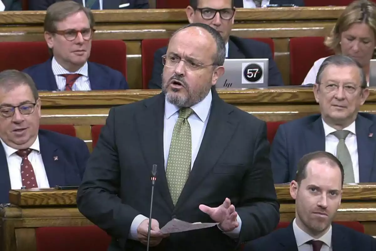 Un home amb vestit i corbata parla en un micròfon en un entorn formal amb diverses persones assegudes darrere seu.